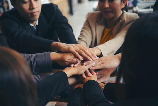 Group of businesspeople joining and holding hands during business meeting at wooden table in office, surrounded by laptops, tablets, documents and charts, showing teamwork, cooperation and unity