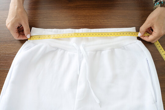 Hands stretch yellow tape measure across white pants waistband laid flat on wooden table, showing garment waist sizing for tailoring, fit checking