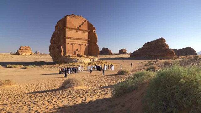 Wide landscape shot of tourists gathering in front of Qasr Al-Farid ancient Nabataean rock-cut tomb under clear blue sky in Hegra, AlUla