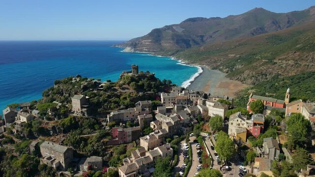 Nonza village aerial drone pan shot with dark Genoese tower and black beach in Cap Corse Corsica. Scenic panoramic rotation over perched cliff town and turquoise sea in 4K stock video