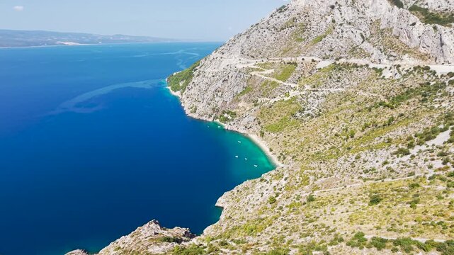 Aerial drone view of Vruja beach a secluded Adriatic bay with crystal-clear turquoise water, rocky cliffs, a coastal road and small boats along the Croatian Dalmatian coast.