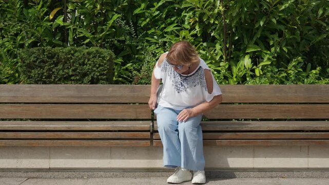 Senior woman sits on a wooden bench in a sunny park to rest. Mature female pensioner relaxes outdoors after a peaceful stroll. Enjoyable retirement lifestyle and comfortable summer recreation.