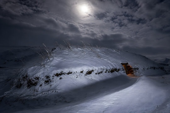 Earth-sheltered mountain refuge almost buried beneath deep snow, with warm light glowing from the entrance on a dramatic moonlit winter night.