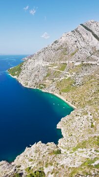 Aerial drone view of Vruja beach a secluded Adriatic bay with crystal-clear turquoise water, rocky cliffs, a coastal road and small boats along the Croatian Dalmatian coast.