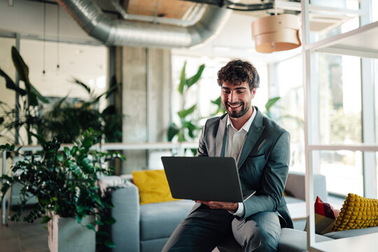 Professional businessman smiling, working remotely on laptop in modern office lounge area, enjoying successful digital work