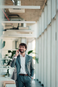Young businessman walking in modern office corridor, talking on phone, smiling and communicating success