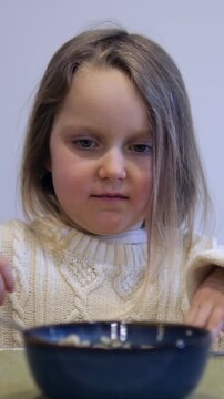Close up of a picky little girl making a disgusted face while eating a bowl of oatmeal with a spoon, highlighting her dislike for the healthy breakfast and her funny expressions