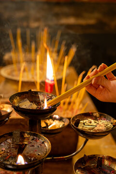An intimate view of a cultural tradition, showing a devotee's hand carefully dipping yellow joss sticks into a burning oil basin to light them for prayer inside a historic temple.