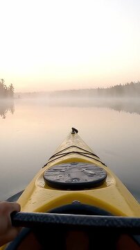Yellow kayak moves across calm lake water as paddler works black double blade paddle. Mist floats above distant forest shoreline while soft morning light creates peaceful outdoor adventure atmosphere.