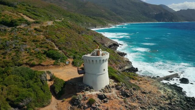 Albo Cap Corse Corsica 4K drone video. Backward fly out reveal of Tour d Albo white Genoese tower on black sand beach with turquoise sea and mountains. Scenic Corsican travel b-roll