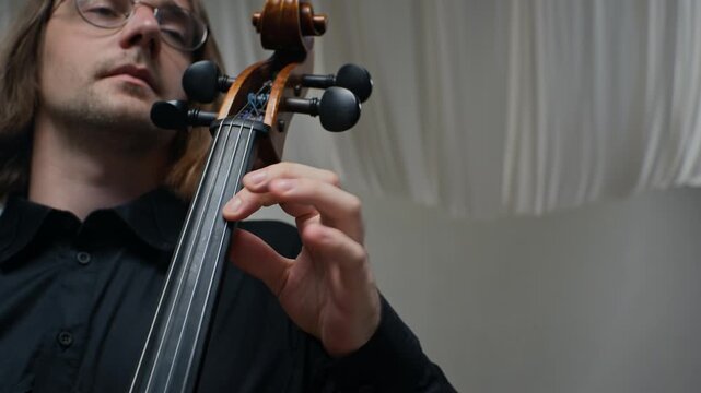 Young adult Caucasian man playing cello, fingers pressing strings on fingerboard in low angle shot. Clip is ideal for music education promos and classical concert advertisements