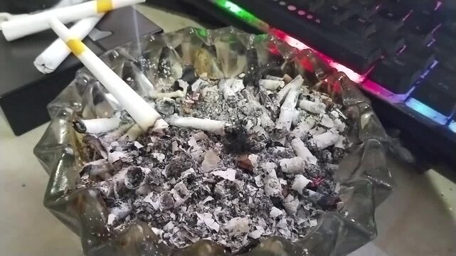Burning Cigarette in Full Glass Ashtray with Ashes Smoke Lighter and Keyboard Background