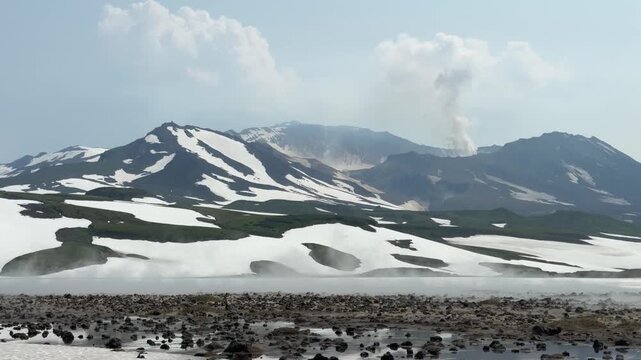 Steam rises from Gorely Volcano crater across melting snowfields and rugged volcanic terrain in Kamchatka. Geothermal activity, volcanic geology, dramatic wilderness, nature tourism, exploration
