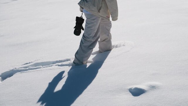 Person walks through deep snow. White pants and jacket stand out. Long shadow stretches behind them. Fresh footprints mark the path. Calm focus shows winter activity. Ideal for travel, winter sports
