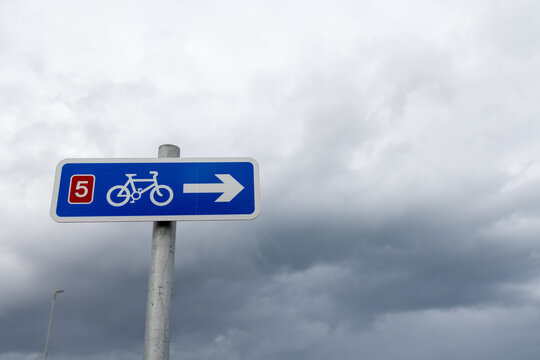 Blue National Cycle Network route 5 direction sign at Penrhyn Bay Wales showing a bicycle symbol and white arrow mounted on a post beneath an expansive cloudy sky near the Welsh coast