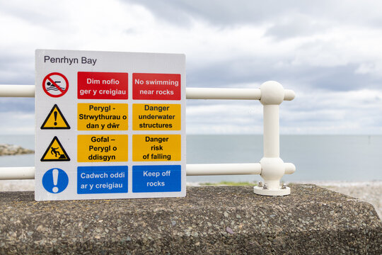 Bilingual coastal safety notice at Penrhyn Bay Wales warning visitors not to swim near rocks and highlighting underwater structures falling risk and instructions to keep off the rocks
