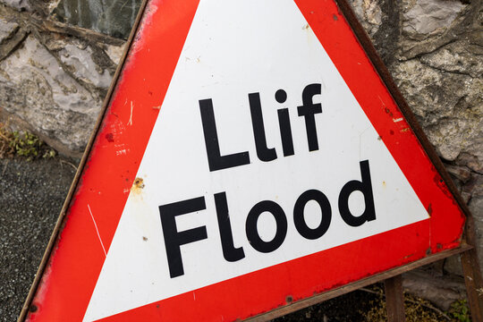 Vintage bilingual Lif Flood warning sign leaning against a stone wall at Penrhyn Bay Wales with bold red triangular border and Welsh and English wording on weathered roadside signage