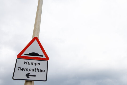 Bilingual road sign for speed humps at Penrhyn Bay Wales showing Humps and Twmpathau with directional arrow beneath a red triangular traffic warning symbol against a cloudy coastal sky