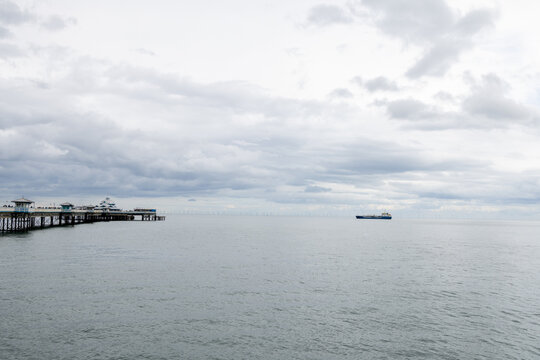 Llandudno Pier extending into the sea in North Wales with a vessel offshore and distant wind turbines visible across the water beneath an expansive cloudy sky on the Welsh coast