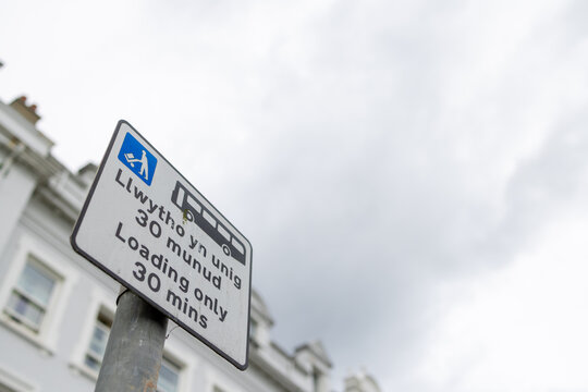 Bilingual loading only road sign in Llandudno North Wales displaying Welsh and English text with a 30 minute restriction and delivery vehicle symbol beside traditional town buildings