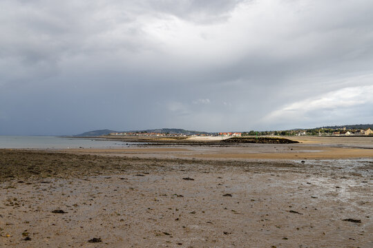 Wide view across Penrhyn Bay North Wales at low tide showing exposed wet sand coastal houses distant hills breakwater and a dramatic cloudy sky over the shoreline
