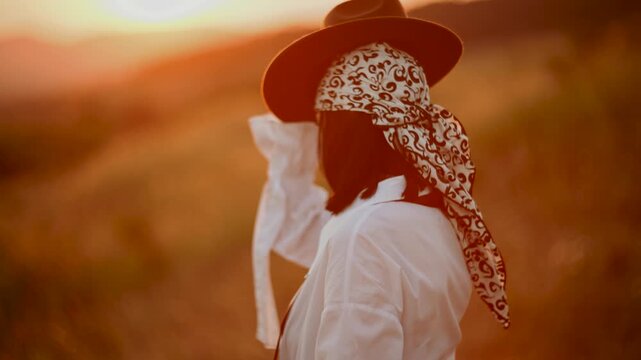 Young woman wearing a patterned headscarf walks through a grassy meadow during a warm golden sunset
