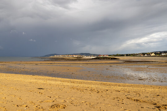 Penrhyn Bay North Wales beach at low tide with broad sandy shoreline shallow water distant coastal homes hills and dark dramatic clouds stretching across the Welsh coast