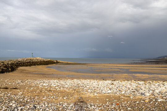 Rocky and sandy shoreline at Penrhyn Bay North Wales during low tide with calm sea exposed beach navigation marker and distant headland beneath a dramatic cloudy sky