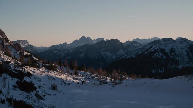 Snowy mountain range bathed in gentle morning light. Clear sky above distant peaks with soft golden hues. Gentle slope covered in fresh snow and scattered trees. Peaceful alpine scene evoking calm