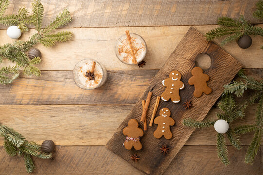 Christmas holiday traditional eggnog on wooden table with gingerbread cookies, cinnamon sticks, star anise, fresh evergreen branches, and brown, and cream-colored ball shaped ornaments.