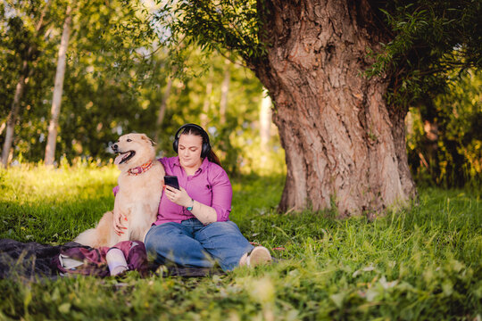 Woman Relaxing with Dog in Park Listening to Music and Using Phone Under a Large Tree