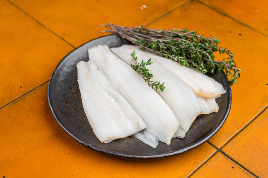 Fresh raw whitefish fillet on metal plate with thyme on bright orange background. Cod, hake or pollock fillet preparation for healthy seafood cooking.