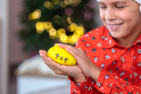 Closeup of smiling child holding yellow squishy toy with cute face, soft hands cradling toy, warm bokeh holiday lights and blurred evergreen tree in background, red patterned shirt suggesting
