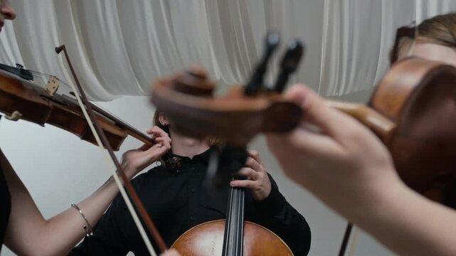 Young adult Caucasian man playing cello alongside young adult Caucasian women playing violins in string trio. Clip is ideal for classical music promotions or artistic collaboration videos