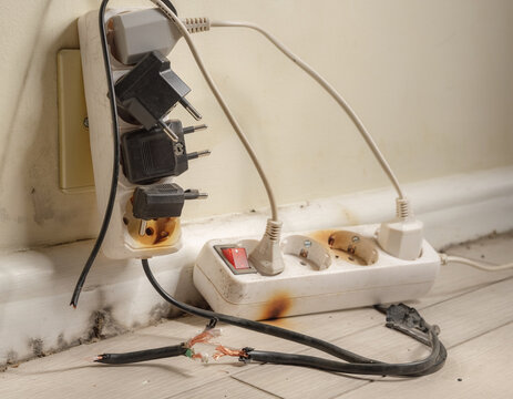 Overloaded electrical wall outlets with multiple power adapters and a damaged, exposed cable, representing unsafe wiring, overheating, fire risk, and household electrical hazards.
