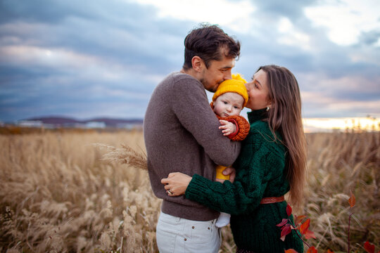 Young parents hugging infant baby girl walking outdoors in fall meadow. Happy loving family kissing daughter enjoying autumn view