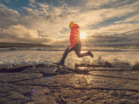 Person in red travel jacket and yellow hat jumps high on rocky shore. Doolin area, Ireland. Sunlight breaks through cloudy sky above. Waves crash around large stones. Popular tourist location.