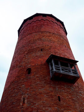 Tall red brick tower with wooden balcony. Ancient structure stands against cloudy sky. Strong, weathered walls suggest historical significance. Ideal for themes like heritage, defense