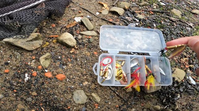 Open tackle box on rocky riverbank filled with colorful inline spinners and metal spoons. Fishing gear assortment for asp (Leuciscus aspius).