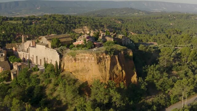 Aerial drone view rotating around the hilltop village of Roussillon, France  surrounded by famous ochre cliffs at sunrise, Provence