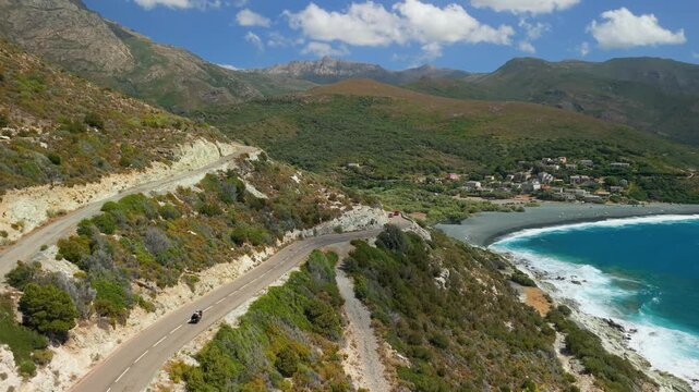 Scenic coastal road drive to Albo black sand beach aerial drone video Corsica France. Mountain serpentine road trip overlooking turquoise sea 4K