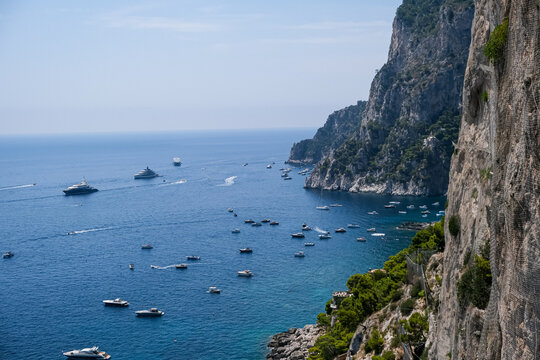 High Angle View of Boats and Yachts by Cliffs in Capri, Italy. A breathtaking, high-angle perspective of the steep, rugged stone cliffs of Capri descending sharply into the calm blue waters of the Med