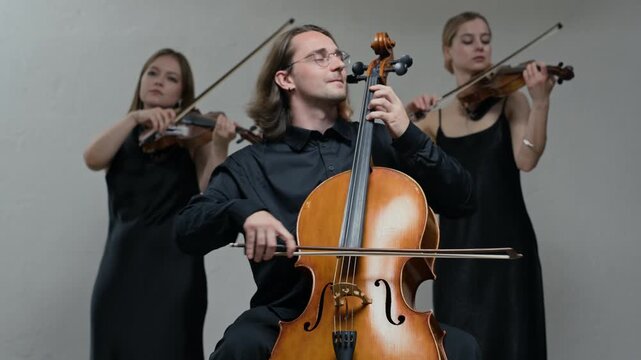 Caucasian young adult male playing cello while two Caucasian young adult female violinists perform in background. Video is perfect for classical music promotions or cultural event advertisements