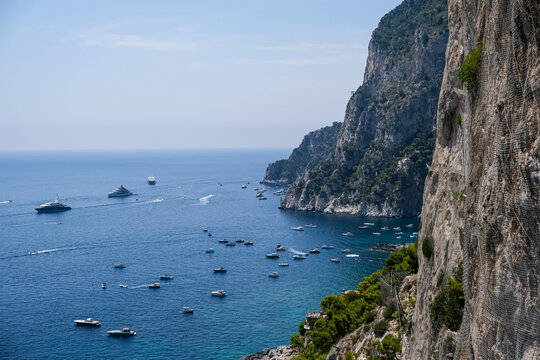 High Angle View of Boats and Yachts by Cliffs in Capri, Italy. A breathtaking, high-angle perspective of the steep, rugged stone cliffs of Capri descending sharply into the calm blue waters of the Med