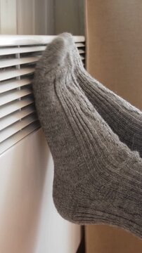 Person feet wearing thick wool socks leaning on radiator grid of electric convector heater. Vertical video. Warming in cold. Energy crisis, high utility bills. Winter house freezing. High heating cost