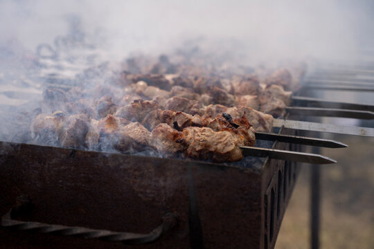 Meat skewers grilling on metal skewers over hot charcoal in outdoor barbecue mangal with rising white smoke.