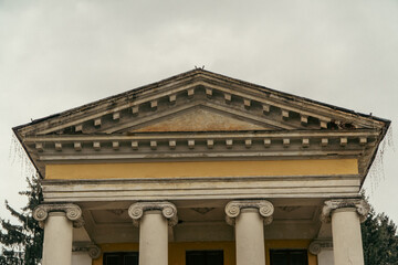 The upper triangular pediment and ionic columns of a historic yellow classical building stand against an overcast sky. © Dmitry