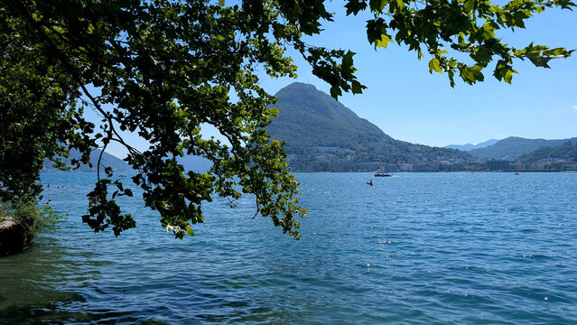 View of Lugano and Mount Bre across Lake Lugano framed by green trees. 