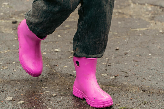 A child wearing dark muddy pants and bright pink rubber boots walks across a rough wet asphalt surface.