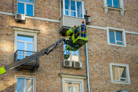 Moscow, August 28 2026, Brick building with multiple windows and air conditioning units. Worker in green lift platform repairs or installs something high up. He wears a helmet and safety gear, focused